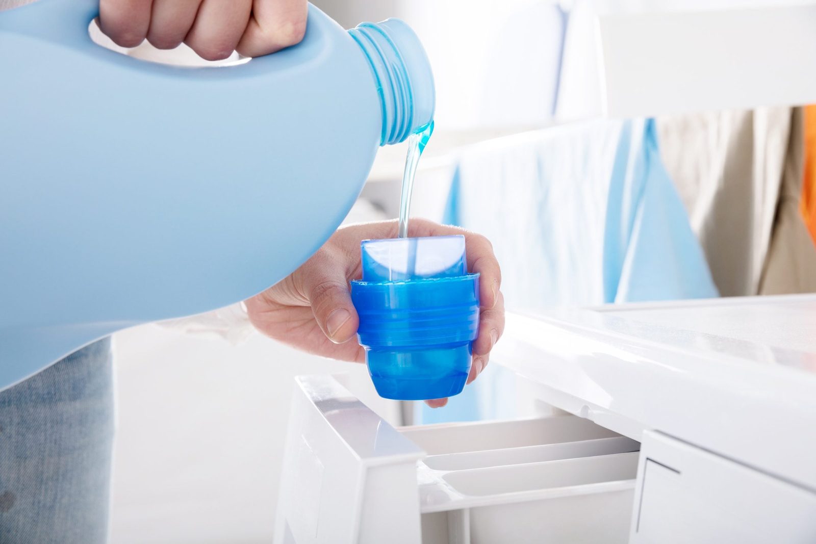 A person pours blue liquid detergent from a jug into a blue measuring cup near a washing machine, surrounded by clean laundry.