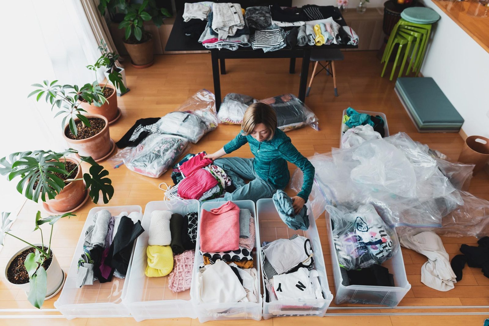 A woman organizes folded clothes into clear bins on a wooden floor, surrounded by plants and additional clothing piles in a bright room.