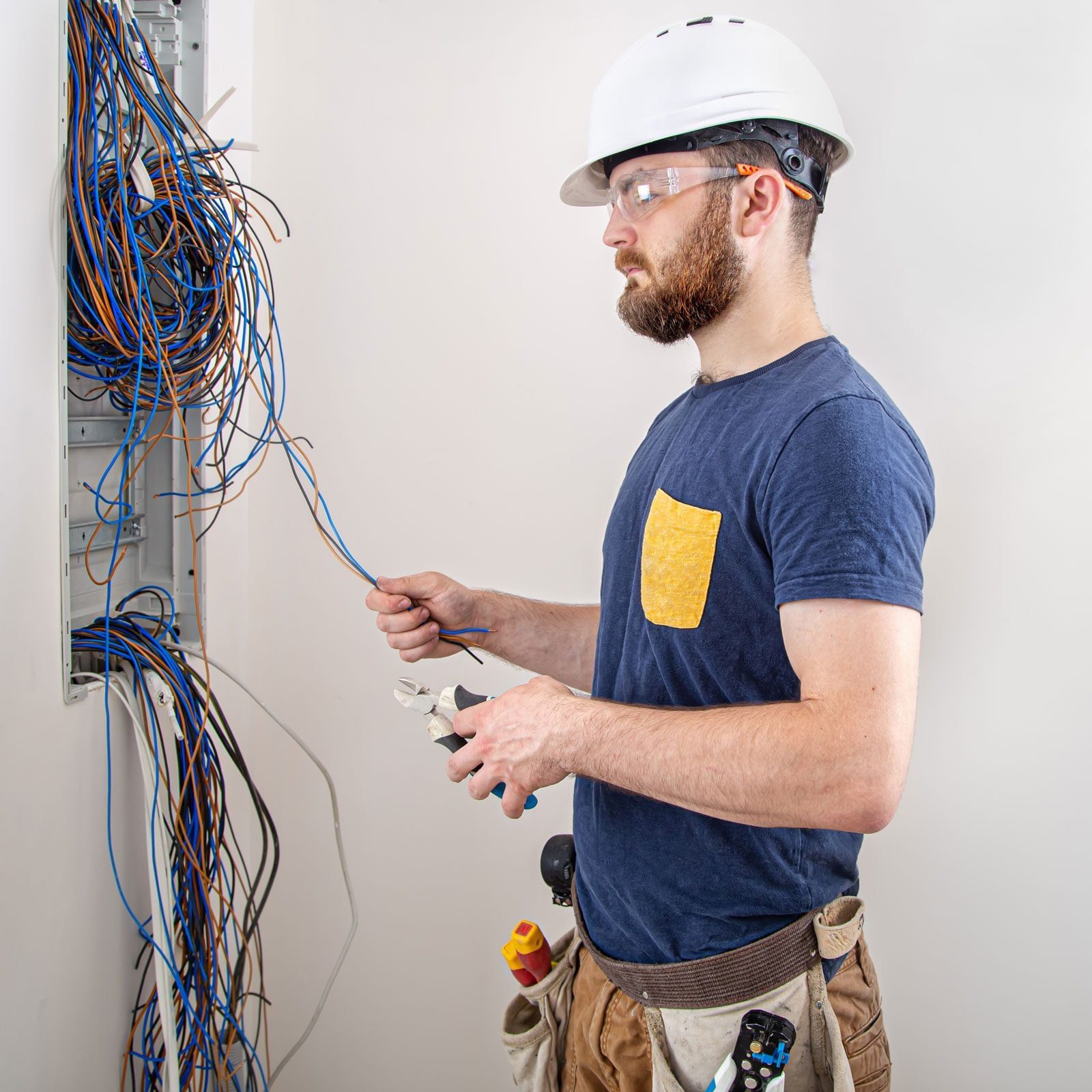 A man in a hard hat and safety glasses works with electrical wires using pliers, while surrounded by a tangle of colorful cables in a neutra