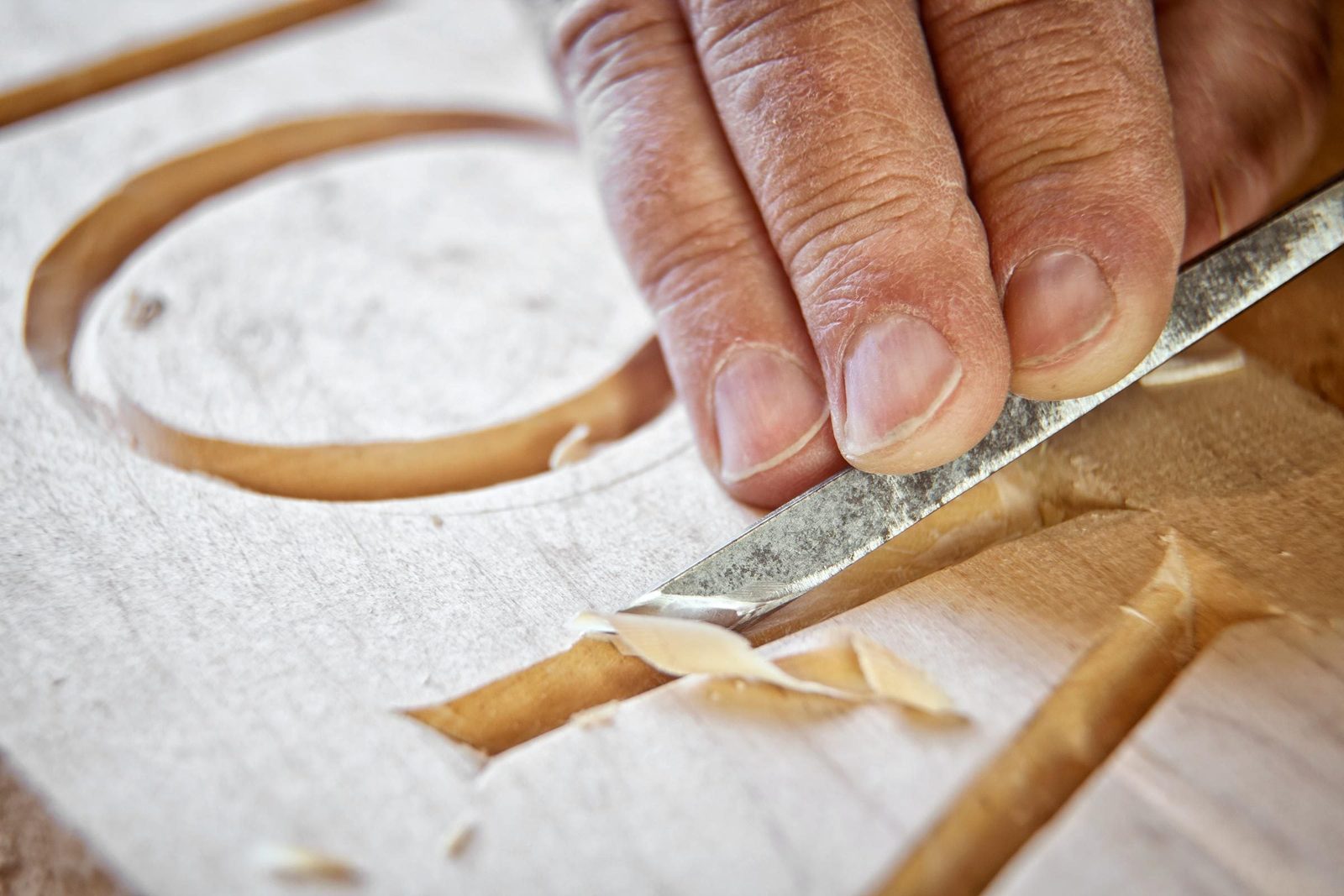 A hand uses a carving tool to shape wood, removing thin shavings from a letter design on a smooth wooden surface.