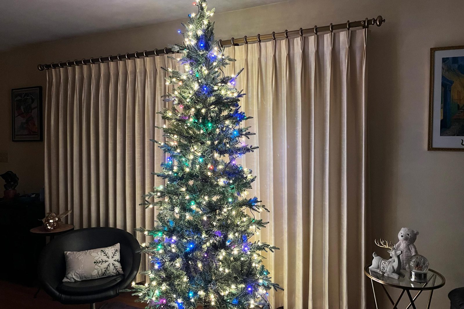 A Christmas tree decorated with multicolored lights stands in front of cream-colored curtains. To the right is a small table with decorative figurines and to the left is a black