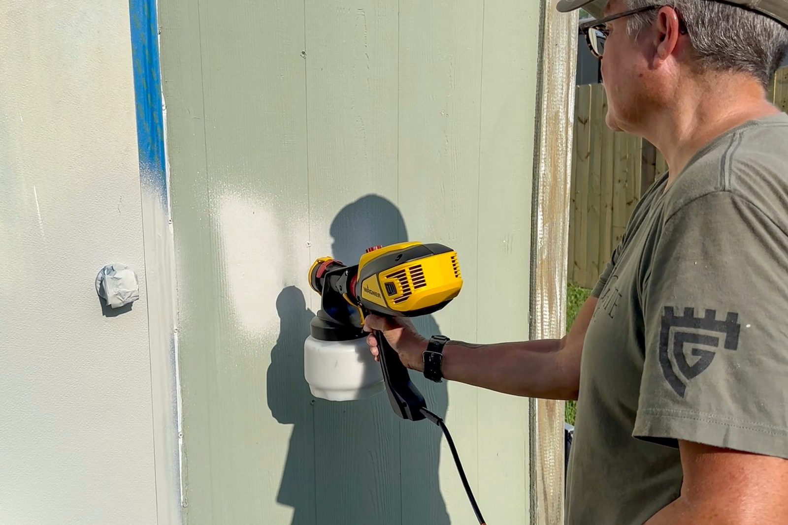A person wearing a beige cap, gray t-shirt, and glasses uses a yellow and black paint sprayer to paint a green vertical surface. The person is standing outdoors next to a wooden