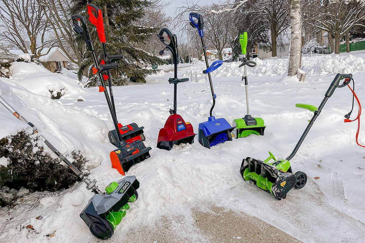 Several electric snow shovels and snow blowers of different colors are lined up on a snow-covered driveway, with trees and houses visible in the snowy background.