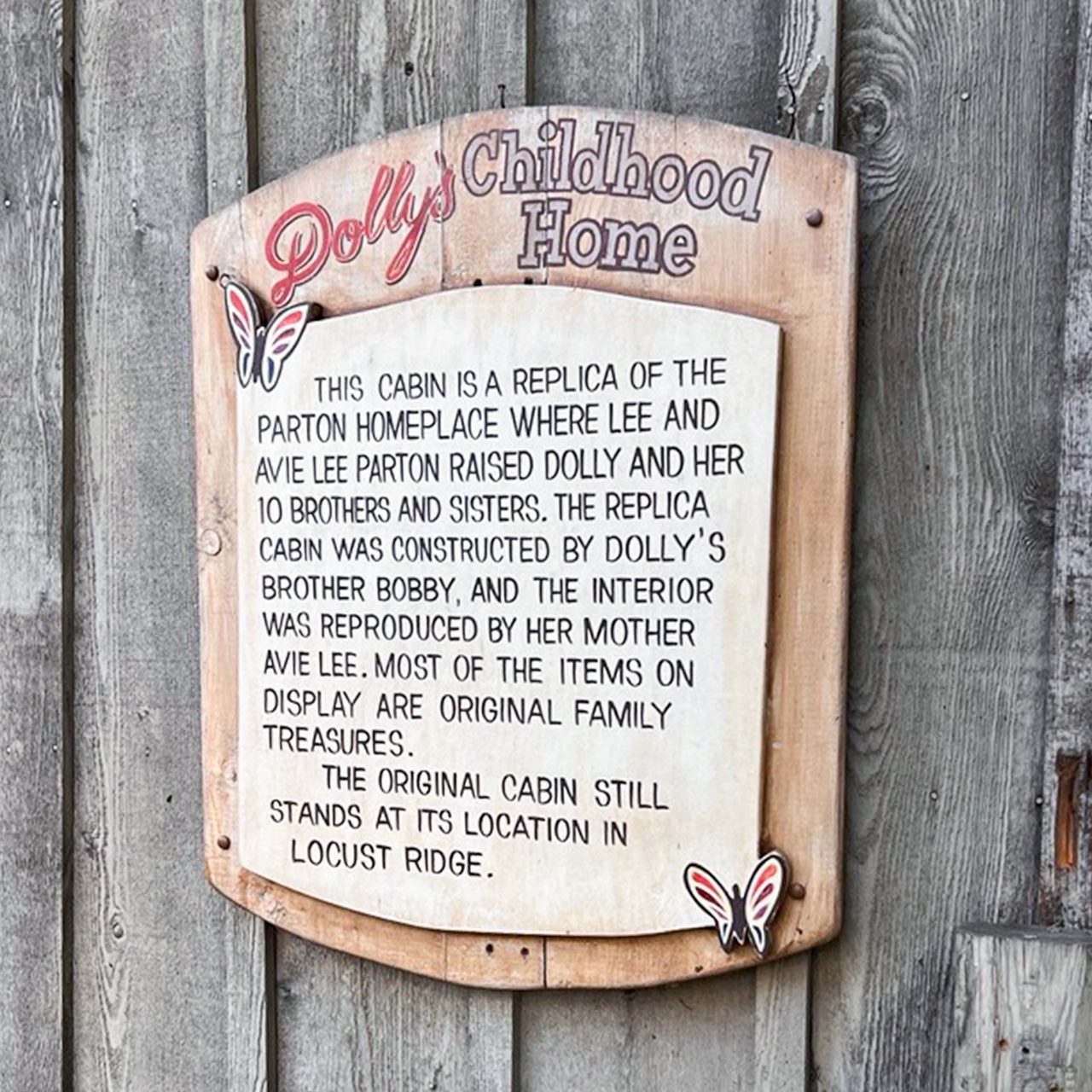 Wooden sign reads: “Dolly’s Childhood Home. This cabin is a replica of the Parton home where Dolly Parton and her family lived, featuring original family treasures. The original
