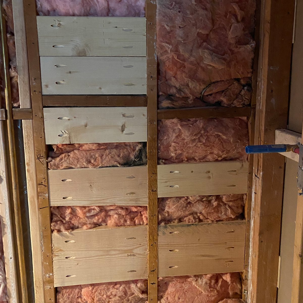 Wooden wall framing with insulation material in pink, showing horizontal wooden boards and a blue pipe.