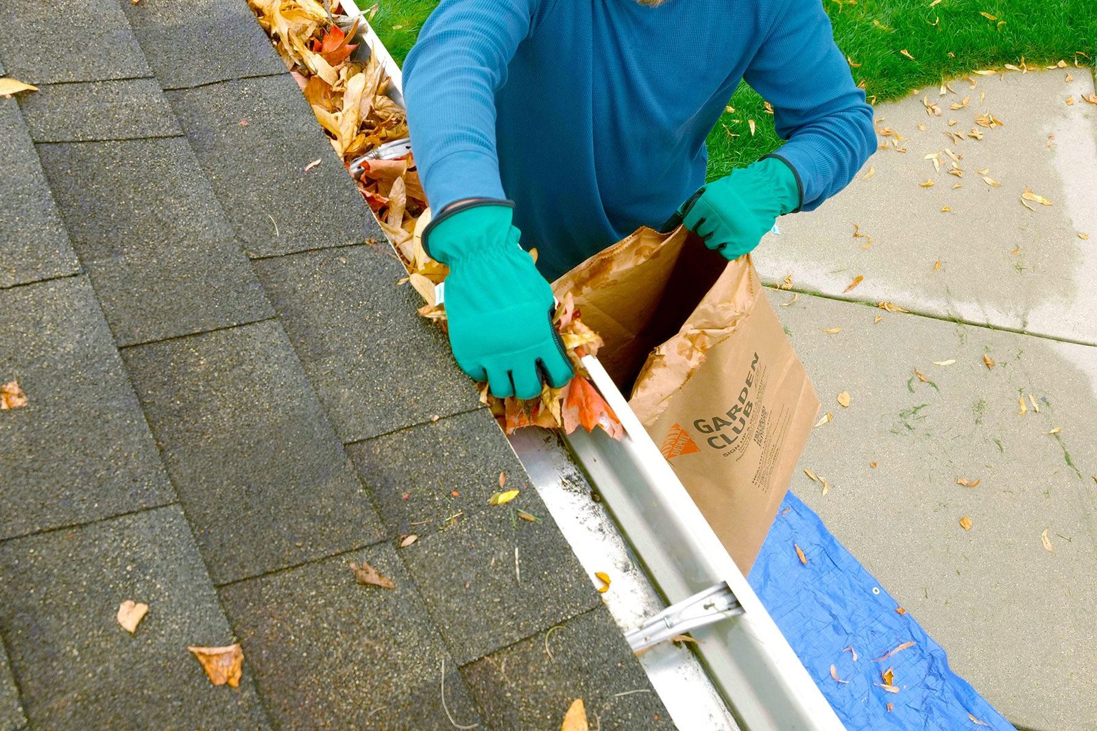 A person wearing gloves removes leaves from a gutter into a paper bag while standing on a roof, surrounded by fallen leaves and a blue tarp below.