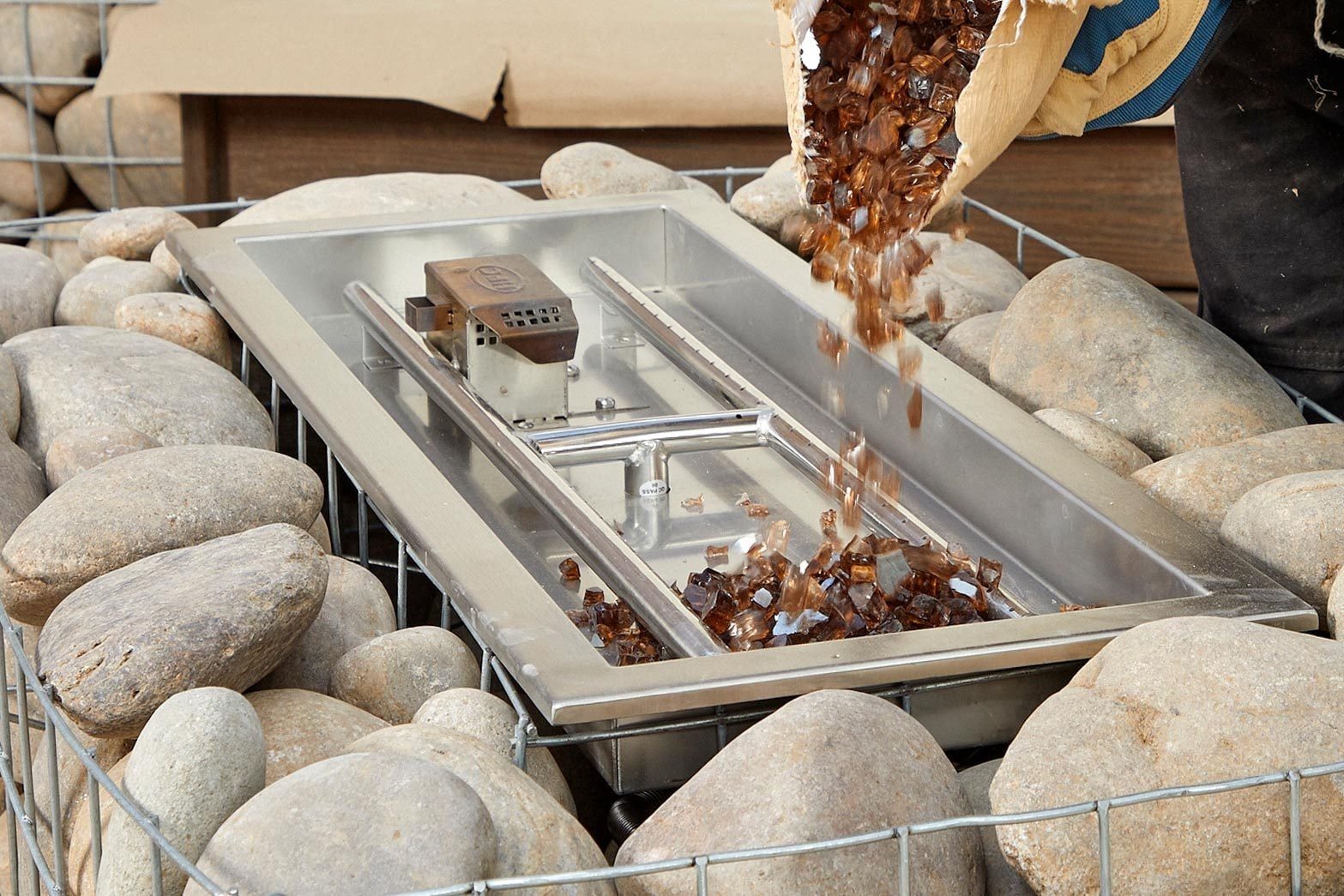 A person is pouring brown glass pebbles into a rectangular metal fire pit surrounded by smooth rocks within a wire frame.