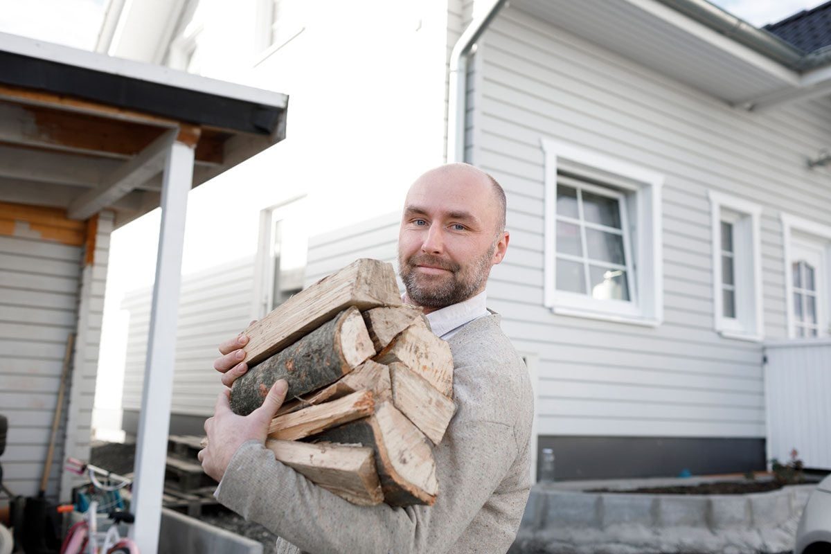 A man carries a stack of firewood while smiling. He stands outside a light-colored house with several windows, surrounded by a patio area.