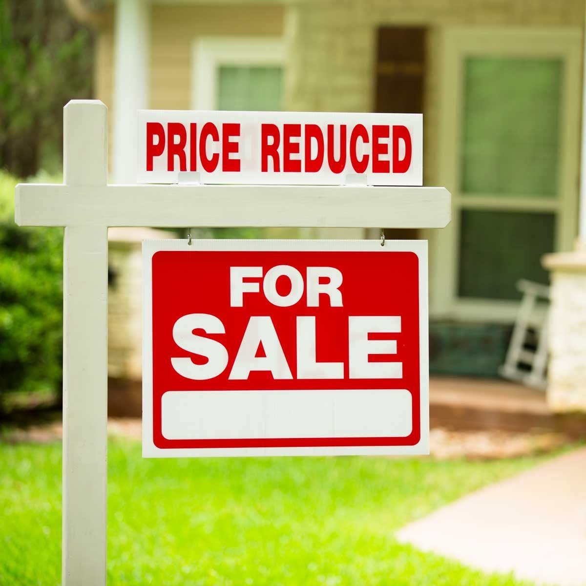 A sign stands in a yard, indicating a home is for sale and the price has been reduced, with a house visible in the background.