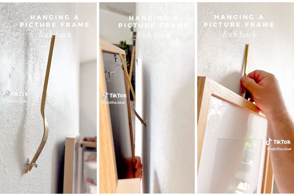 A fork is wedged into a wall as a person positions a picture frame. The setting is a well-lit room with soft decor.