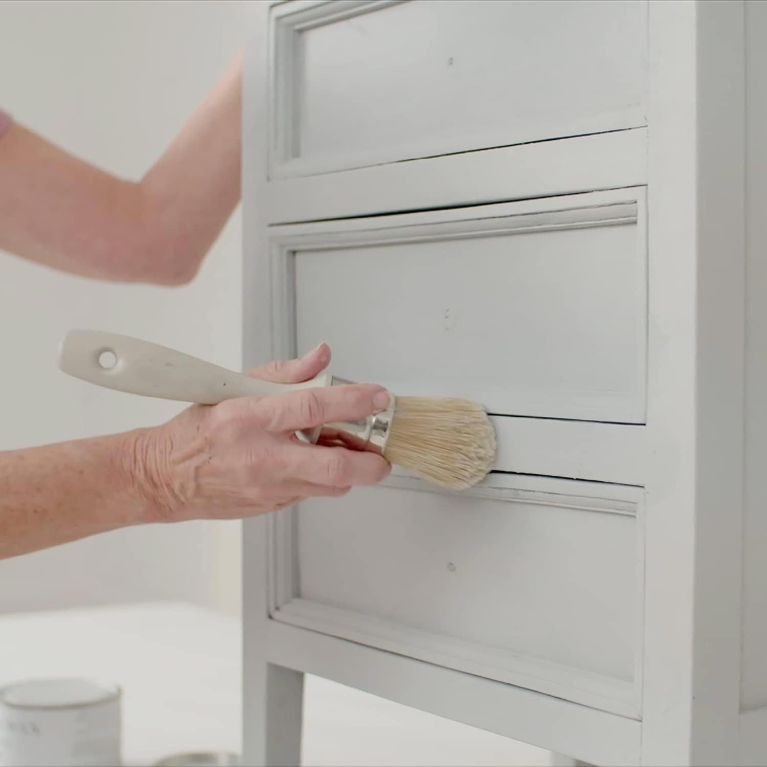 A hand holds a paintbrush, applying paint to the edges of a white-painted dresser in a bright, minimally decorated room.