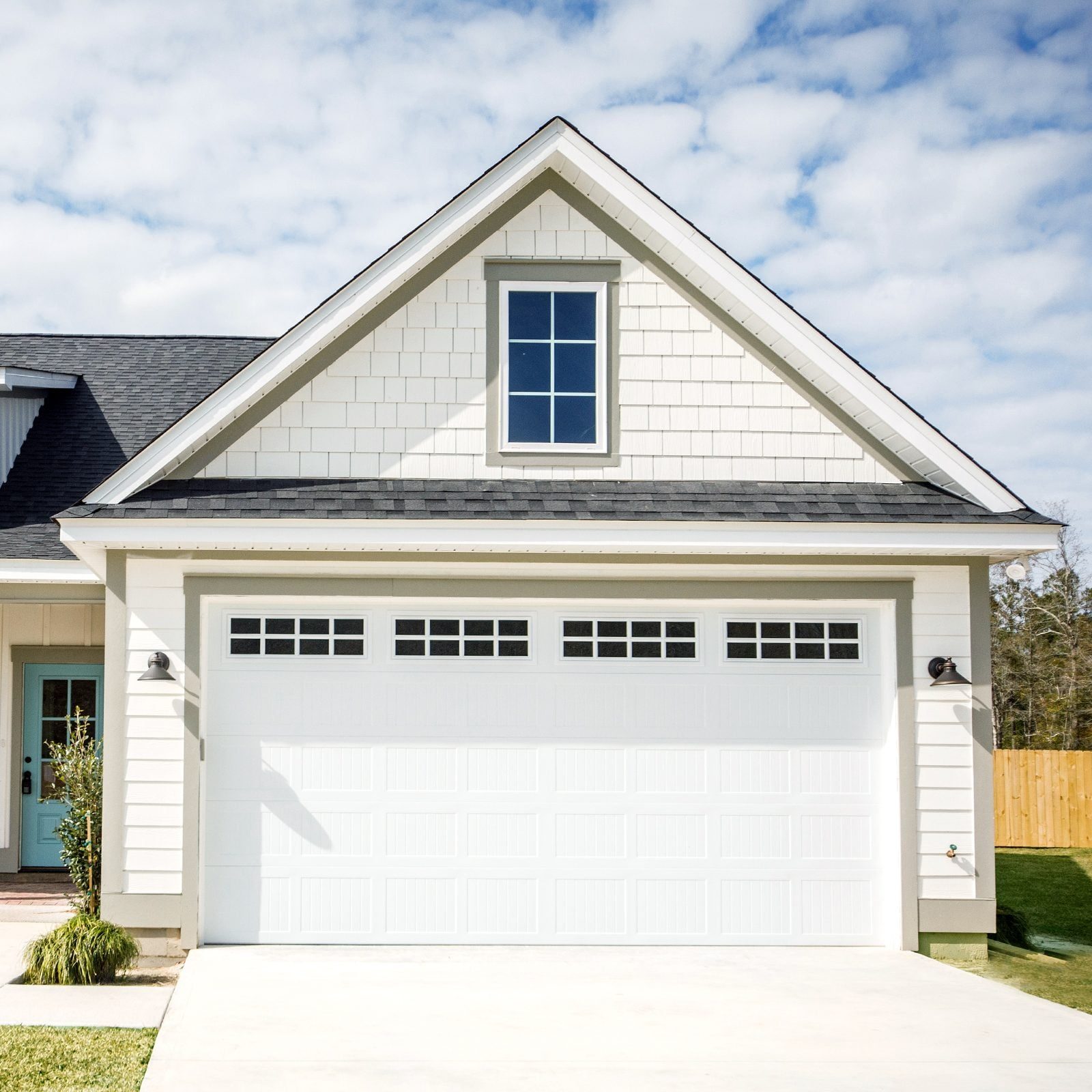 A garage door with multiple windows opens outward, set against a light-colored house. The roofline is triangular, under a partly cloudy sky.