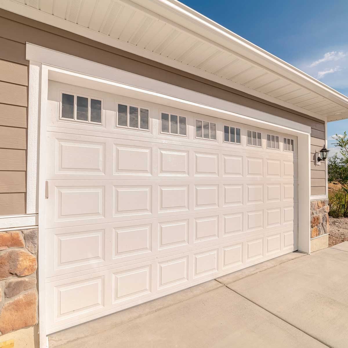 A white garage door stands closed, featuring rectangular panels and small windows at the top, beside a light fixture on a beige house exteri