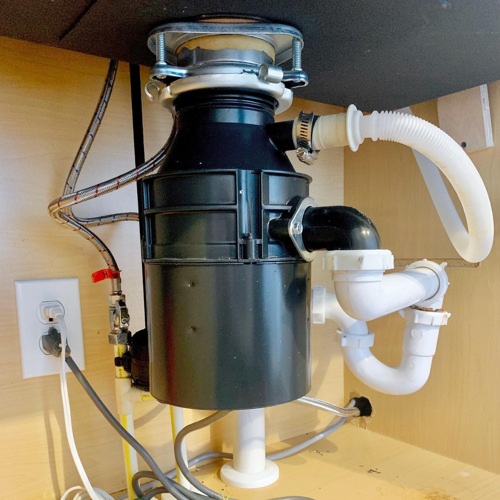 A black garbage disposal unit is installed under a kitchen sink, connected to pipes and power source, in a wooden cabinet environment.