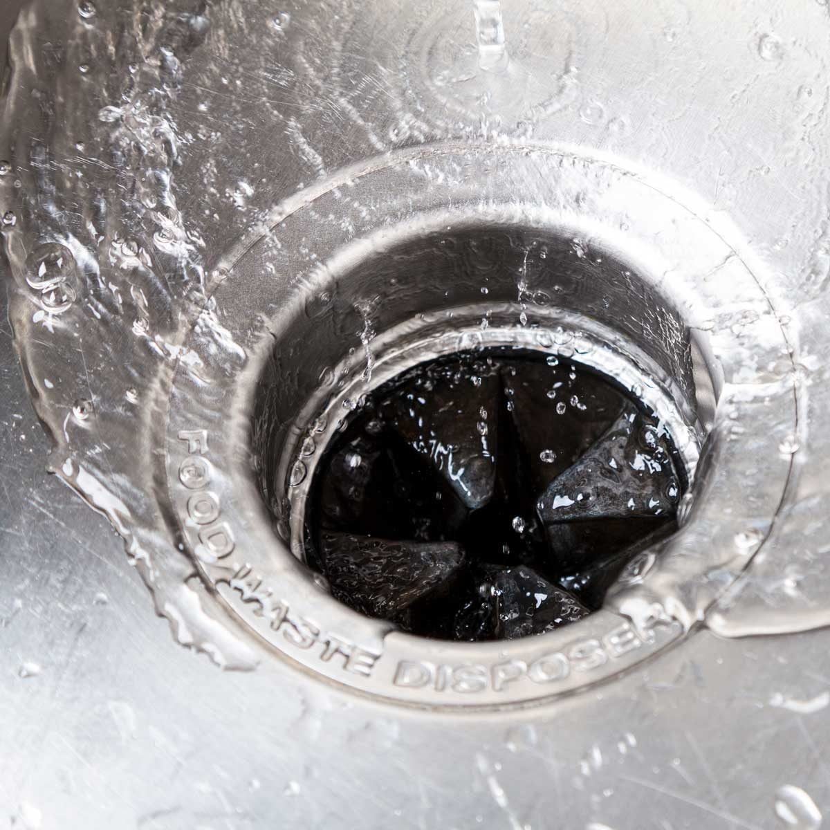 Water cascades into a stainless steel sink's food waste disposal, creating splashes as it rushes toward the dark interior mechanism below.