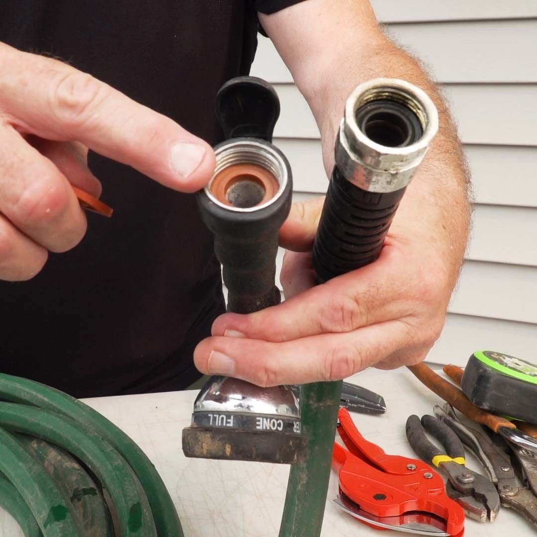 A hand points at a hose nozzle's connection while holding an additional piece. Tools are scattered on a table, with a background of wooden s