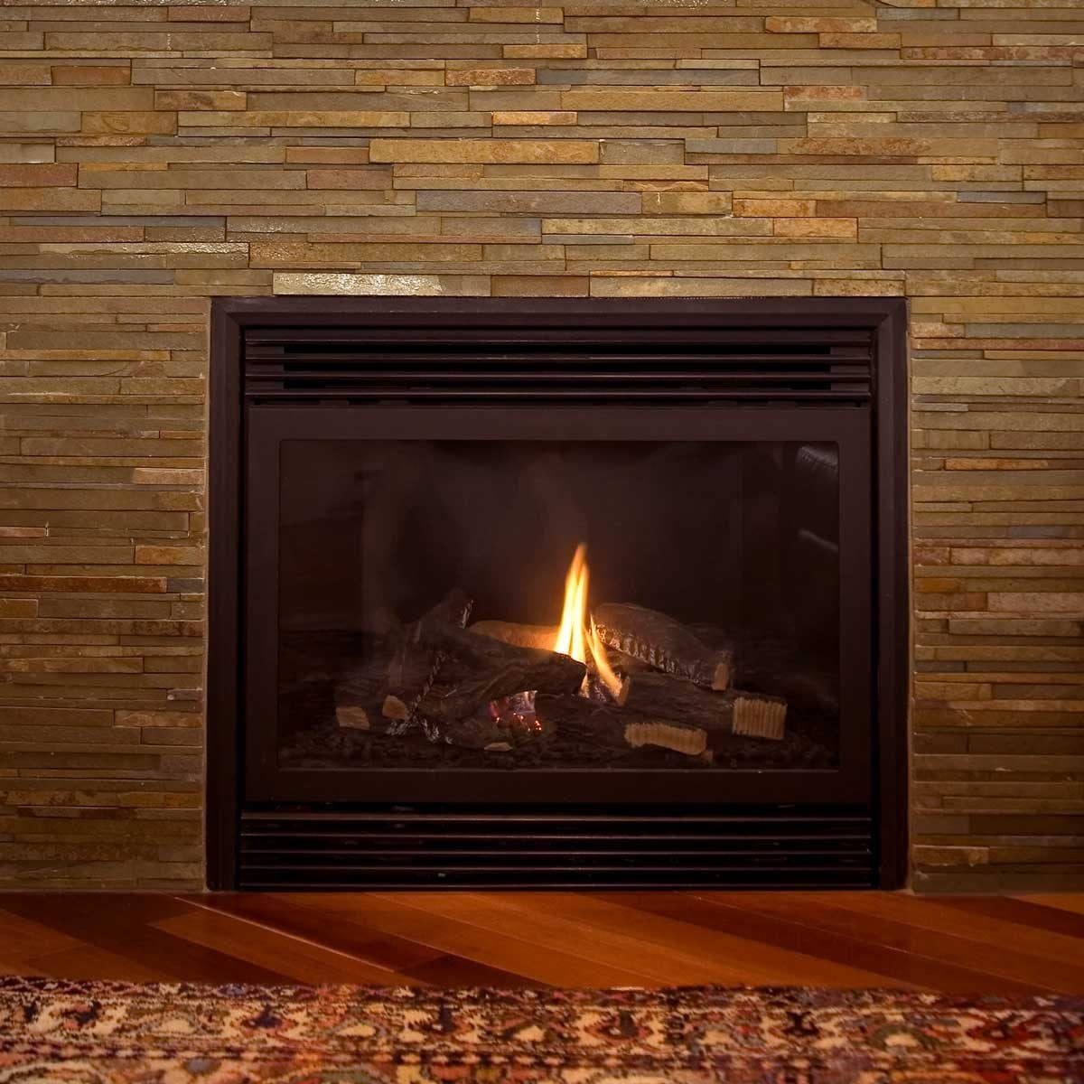 A fireplace with a small flame burns on the logs, surrounded by a textured stone wall and a wooden floor with a patterned rug in front.
