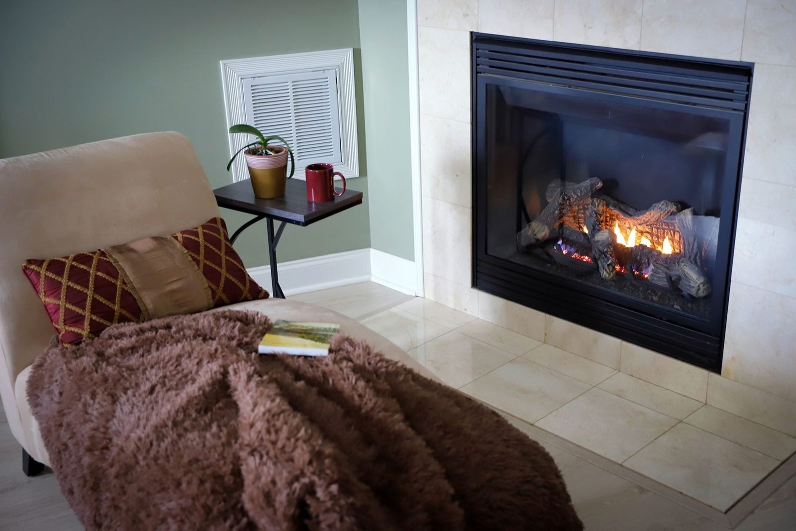 A cozy chair with a brown blanket sits beside a warm fireplace, accompanied by a side table holding a mug and a potted plant.