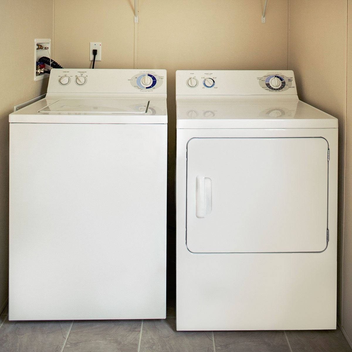 Two white laundry appliances, a washing machine and dryer, sit side-by-side against a neutral wall in a tiled laundry room. Controls are vis