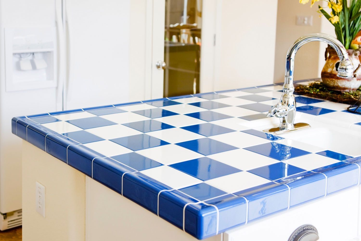 A blue and white tiled countertop features a chrome faucet positioned above a sink, surrounded by a bright kitchen area with visible applian