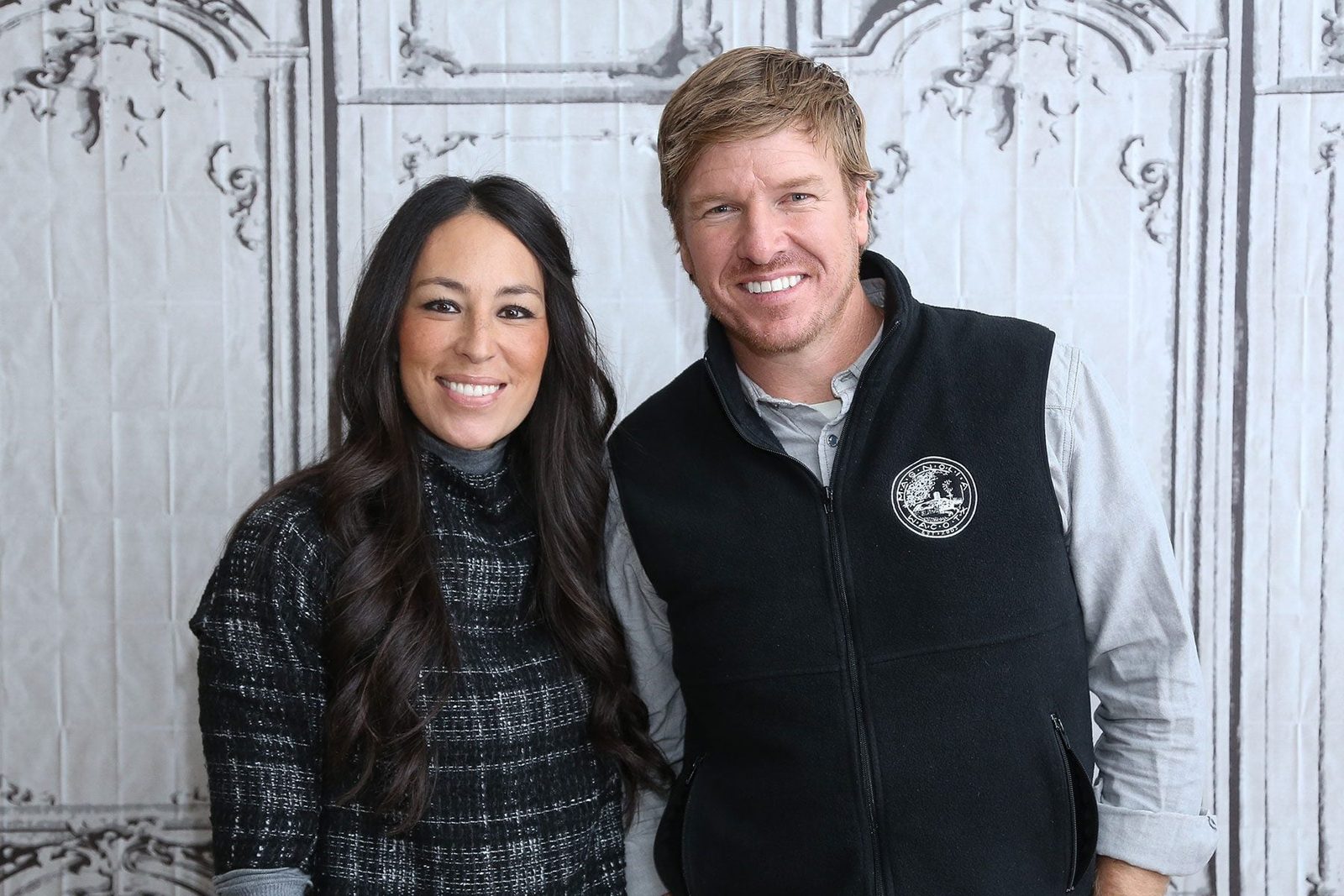 Two smiling individuals stand closely together, dressed casually, in front of a decorative backdrop that suggests a vintage setting, with in