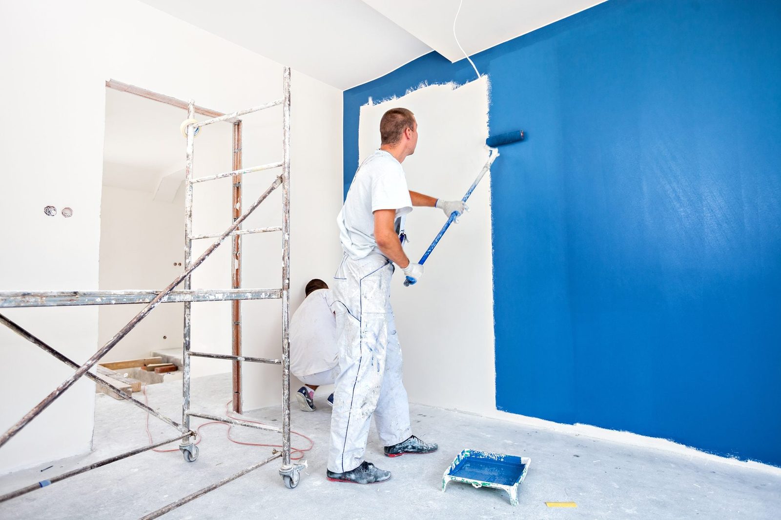 Two people paint a wall; one rolls blue paint while the other works nearby. The room has scaffolding, a ladder, and unfinished surfaces.