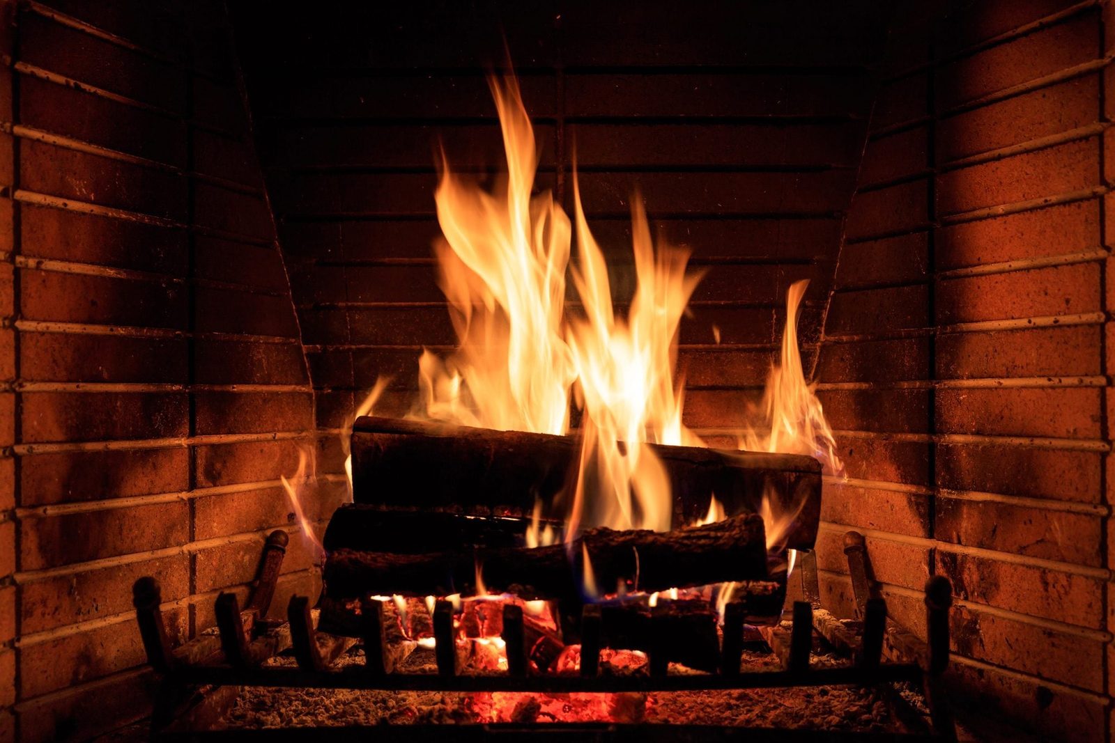 Flames dance over burning logs in a fireplace, illuminating the surrounding brick walls with warm orange hues, creating a cozy atmosphere.