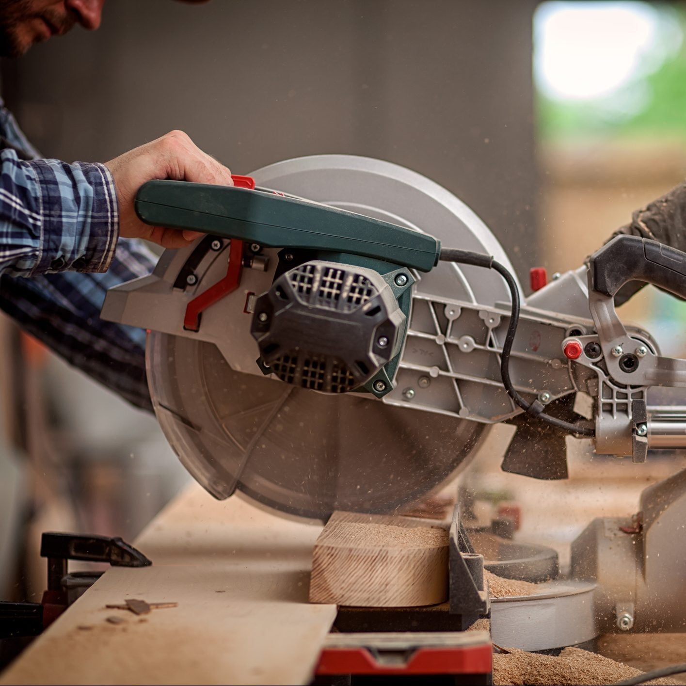 A person operates a power miter saw, cutting a wooden plank, surrounded by sawdust in a workshop setting.