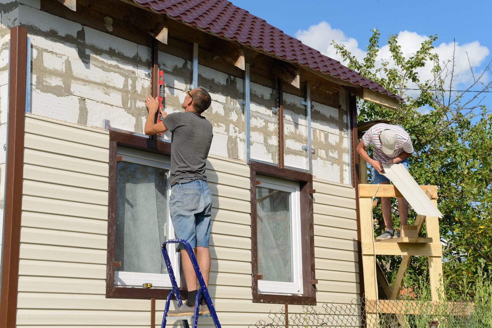 Two men work on a house's exterior; one levels a frame on a ladder while the other installs siding from a wooden platform under a clear sky.
