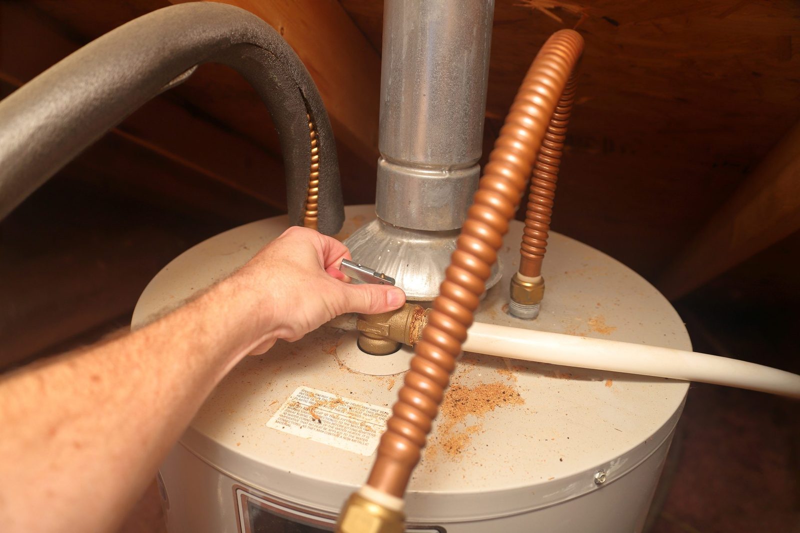 A hand adjusts a valve on a water heater, surrounded by copper pipes and dust, in a dimly lit attic space.