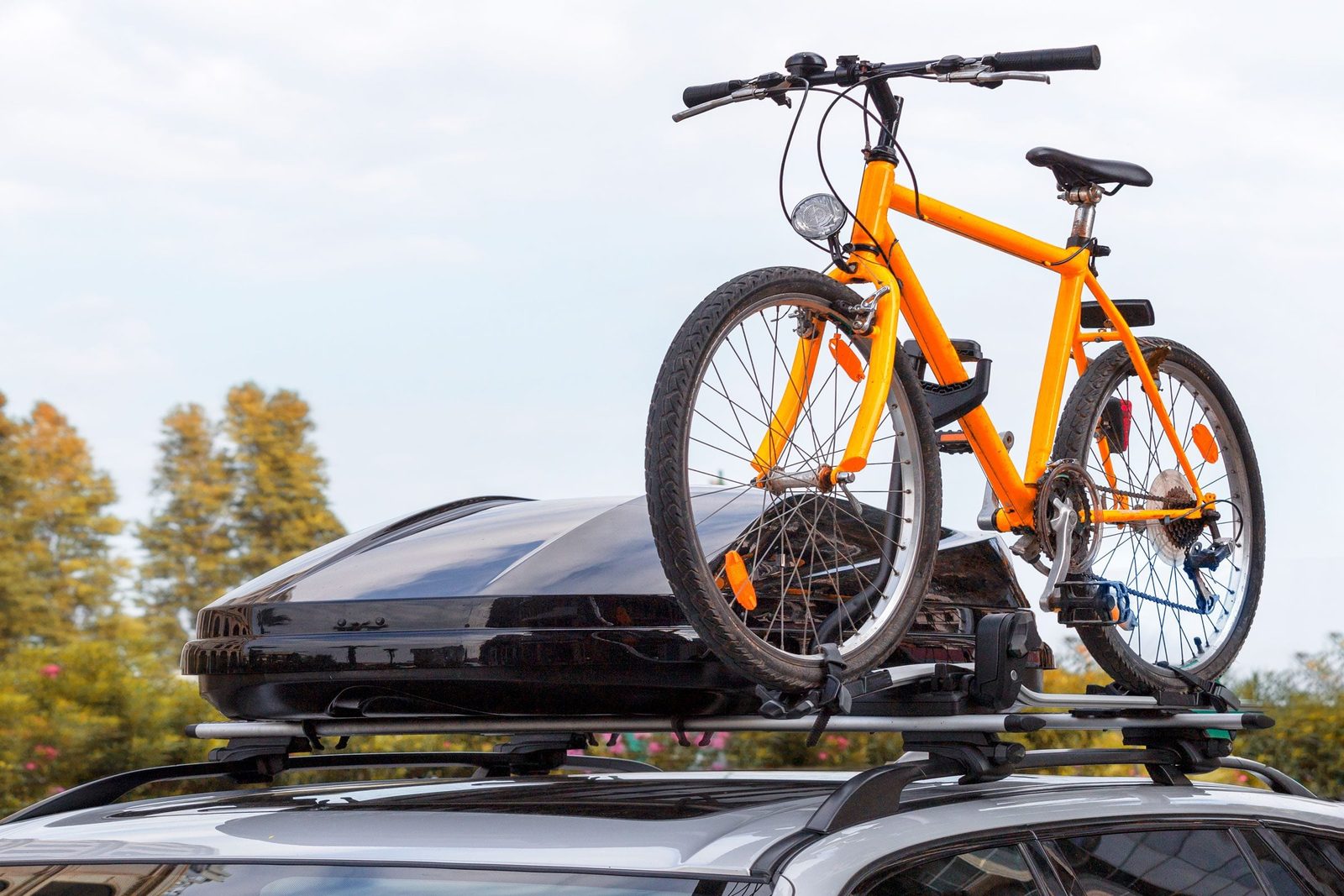 An orange bicycle is mounted on the roof of a car, positioned above a black roof box, set against a backdrop of trees and cloudy sky.
