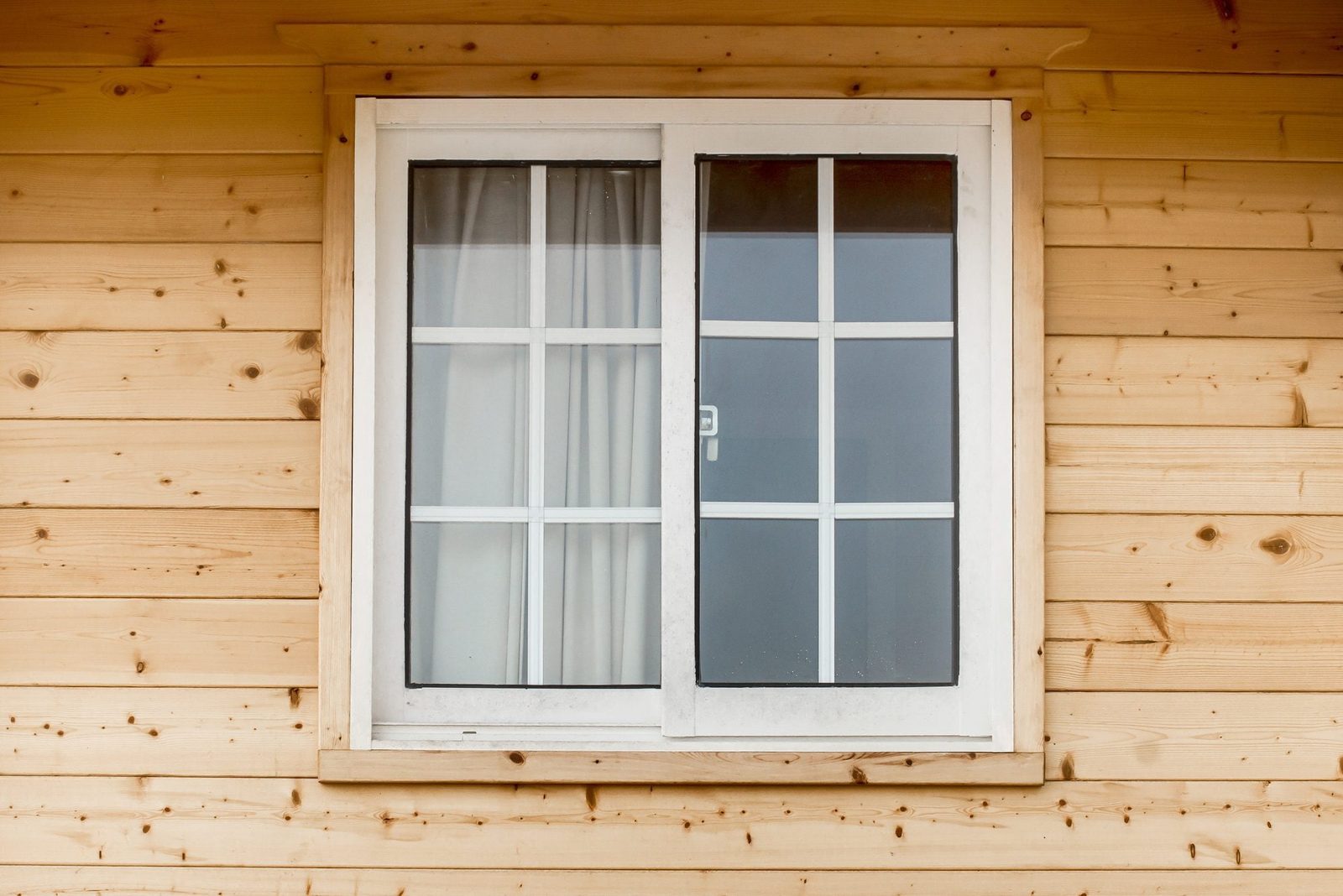 A white window with grid patterns sits on a wooden wall, partially revealing a curtain behind the glass in a cozy setting.