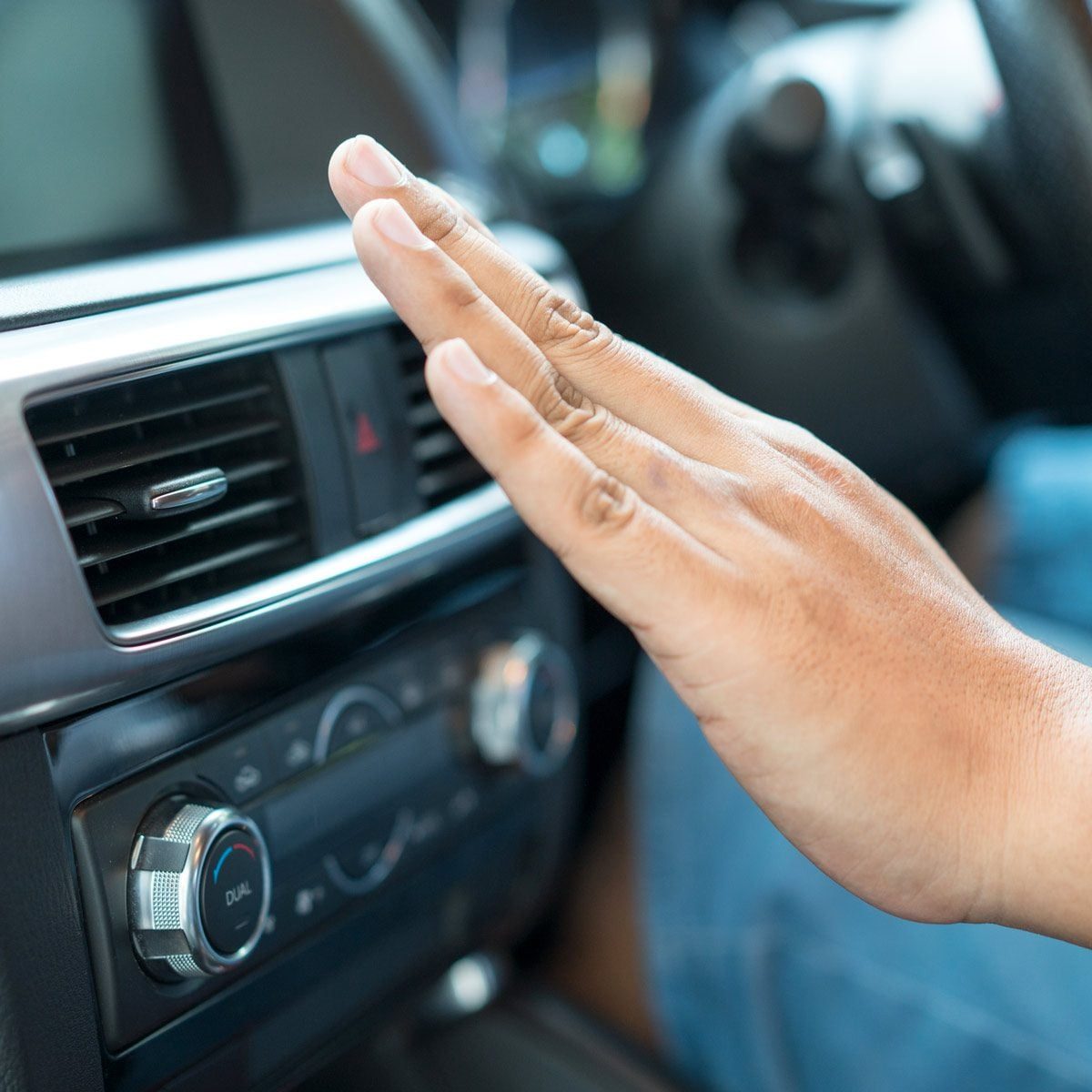A hand is gesturing towards a car's air vent, likely adjusting the climate control, inside a vehicle's interior.