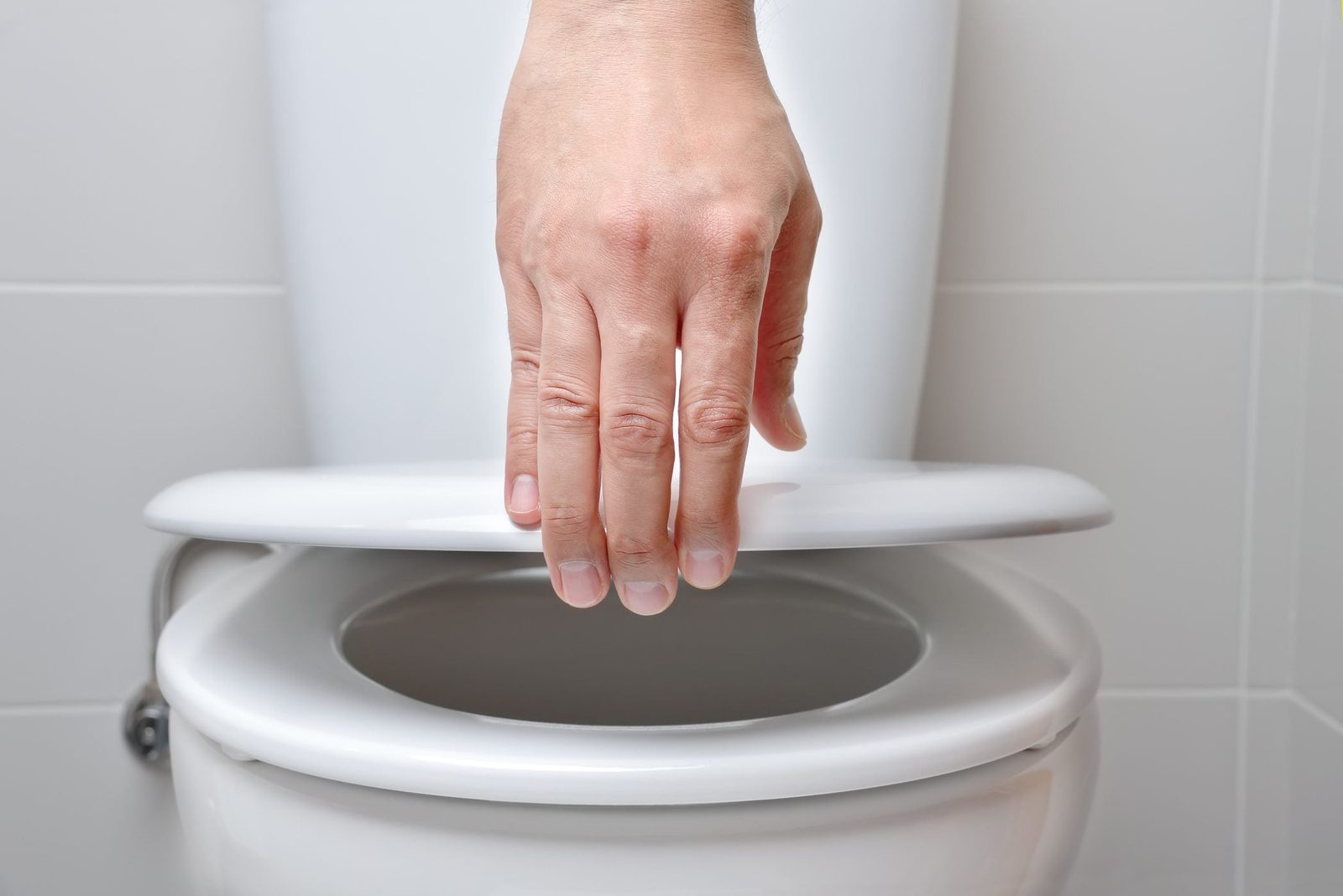A hand lowers a toilet seat into place above a toilet bowl in a clean, bright bathroom.