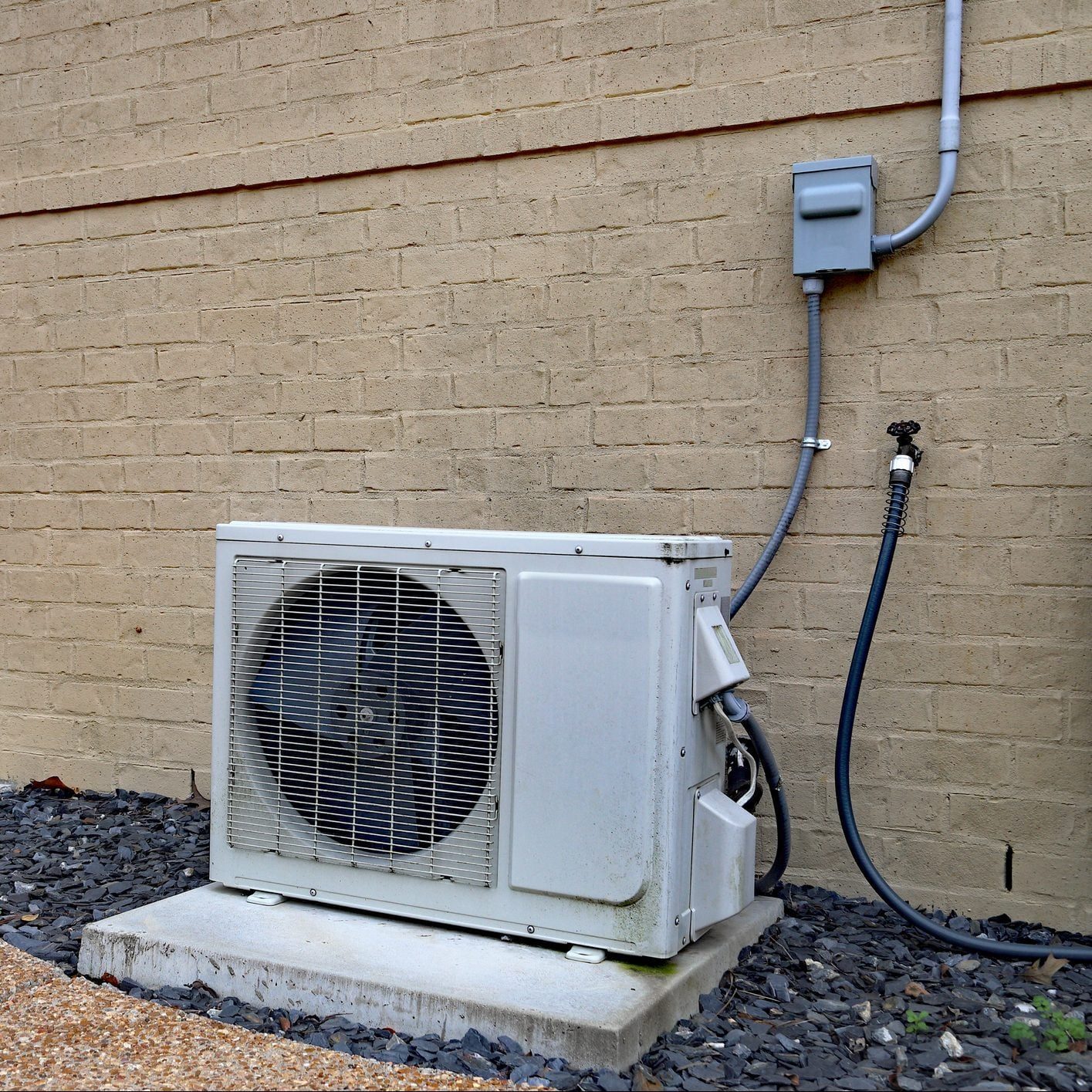 A white air conditioning unit sits on a concrete slab beside a textured beige wall, connected by pipes, with gravel landscaping surrounding