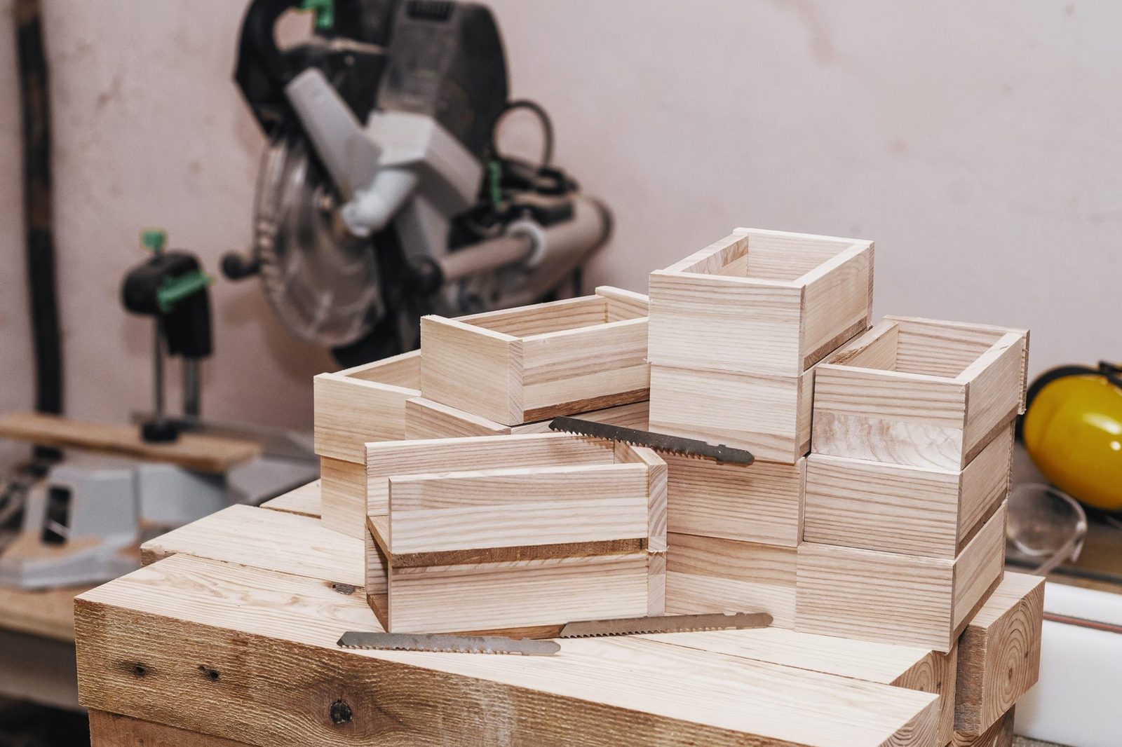 Wooden boxes are stacked on a workbench, while a saw rests beside them in a workshop setting. Tools are visible in the background.