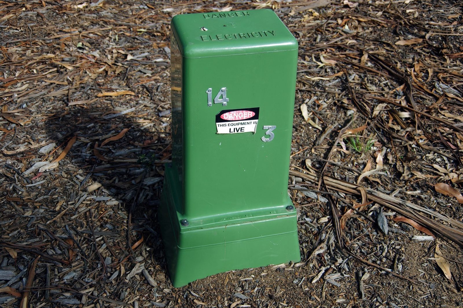 A green electrical box stands on the ground, marked with "Danger: This equipment is live," surrounded by dry leaves and dirt.