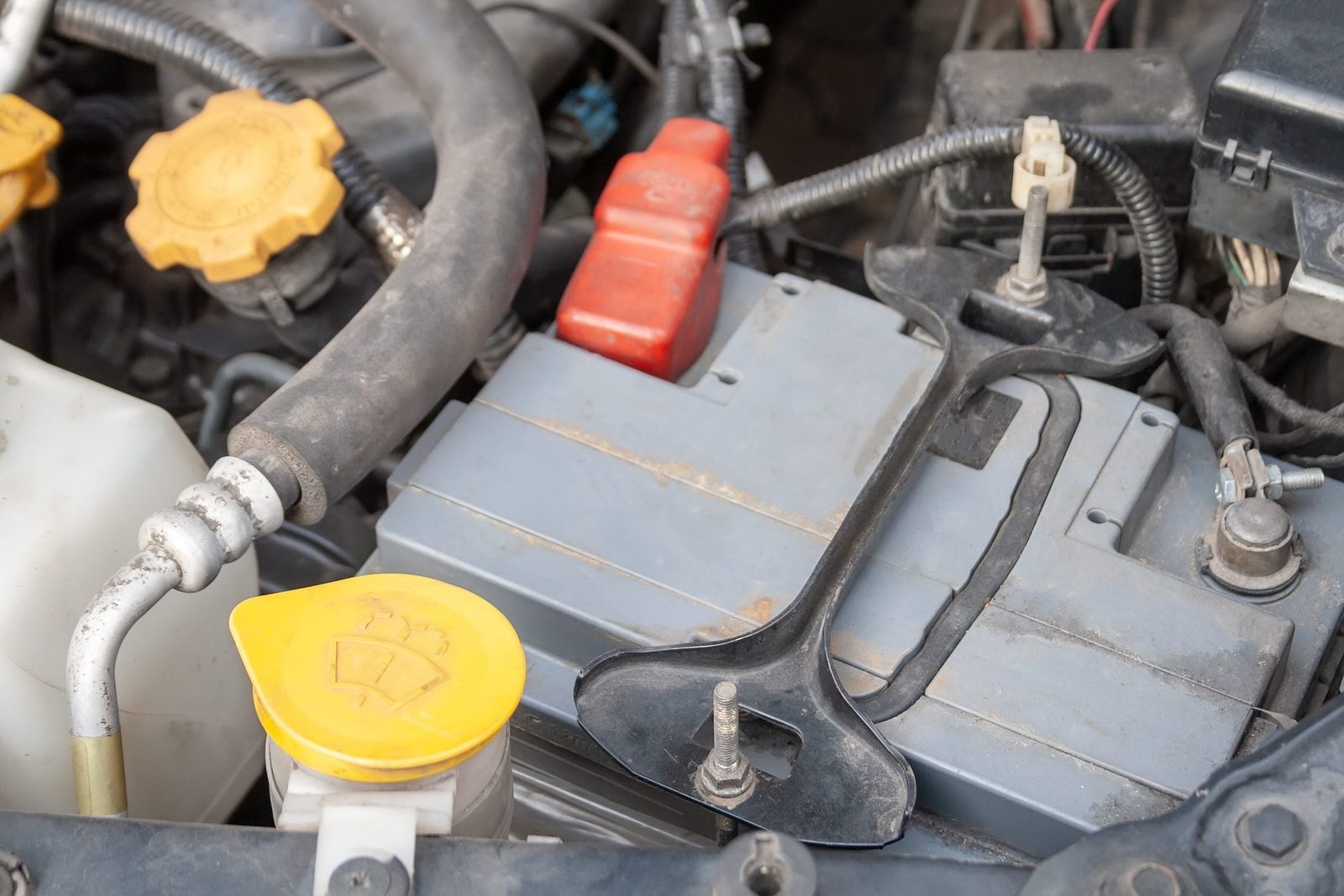 A car battery with connectors and a tool rests in a dusty engine bay, surrounded by hoses and fluid caps, indicating maintenance activities.