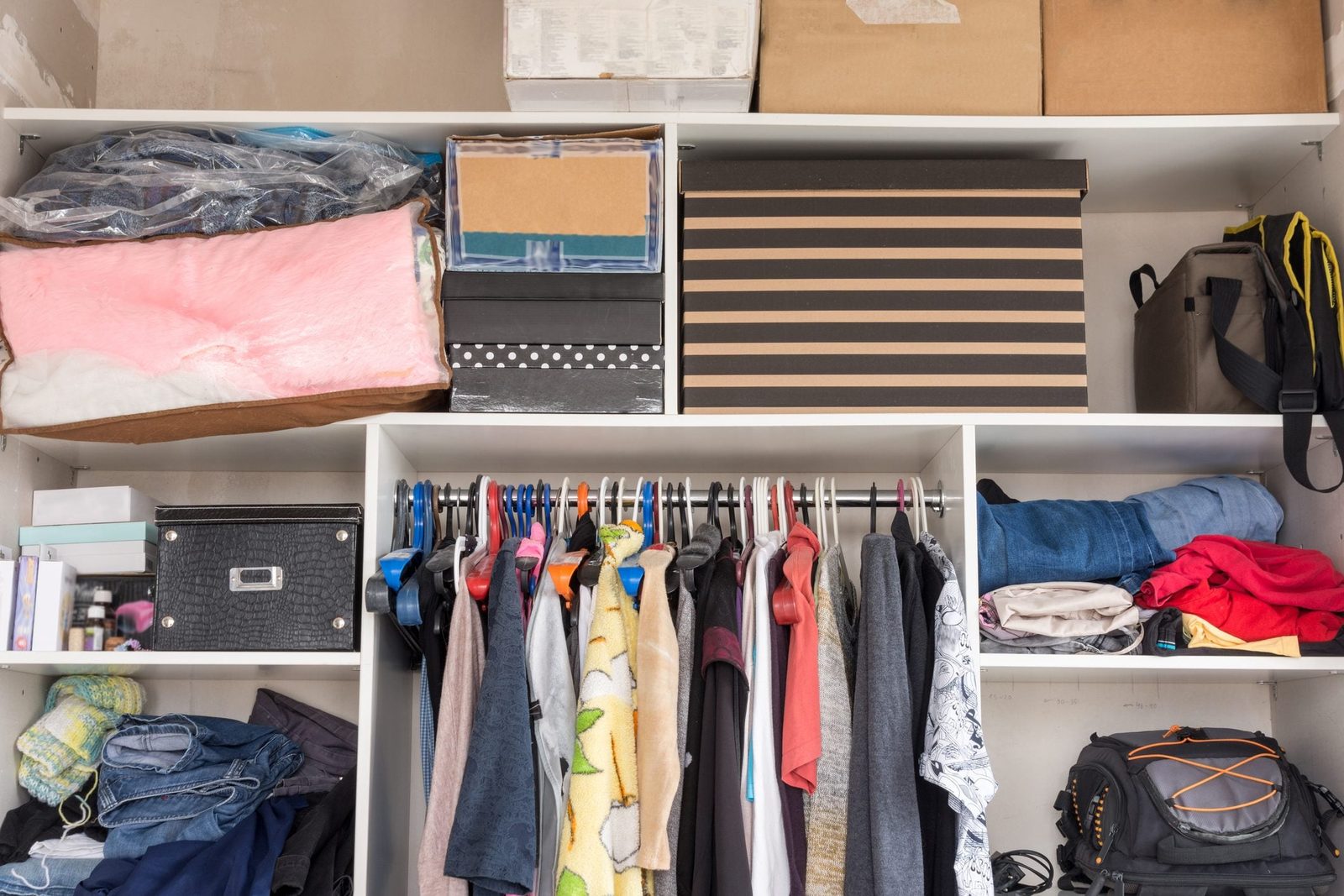 A closet contains various clothes hanging on hangers, organized boxes and bags on shelves, creating a cluttered yet functional storage space