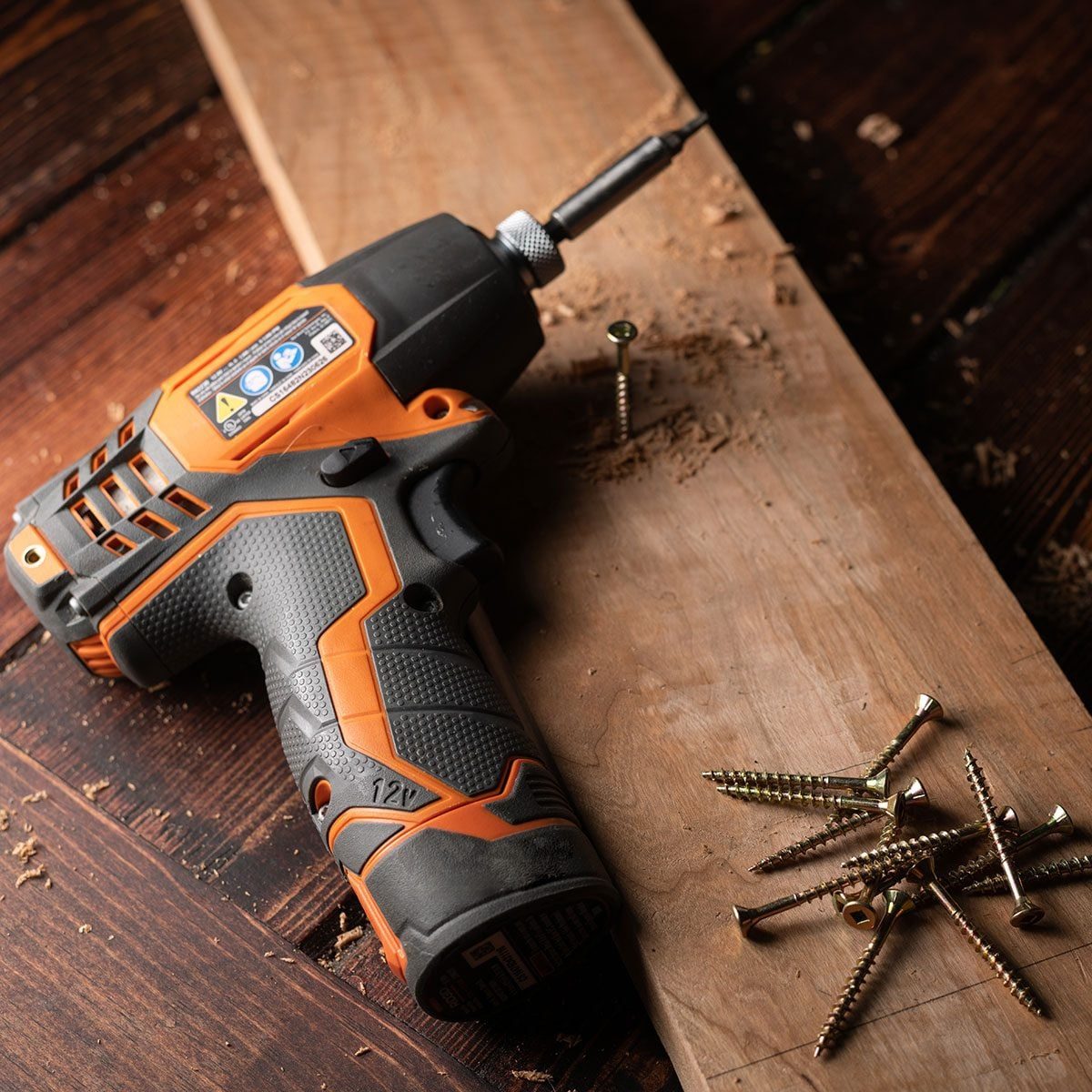 A cordless drill rests on a wooden board, poised to drive screws into the surface, surrounded by scattered screws and wood shavings.