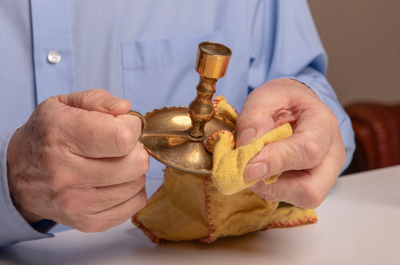A person cleans a brass candlestick with a yellow cloth, seated at a table, with a blurred brown object in the background.