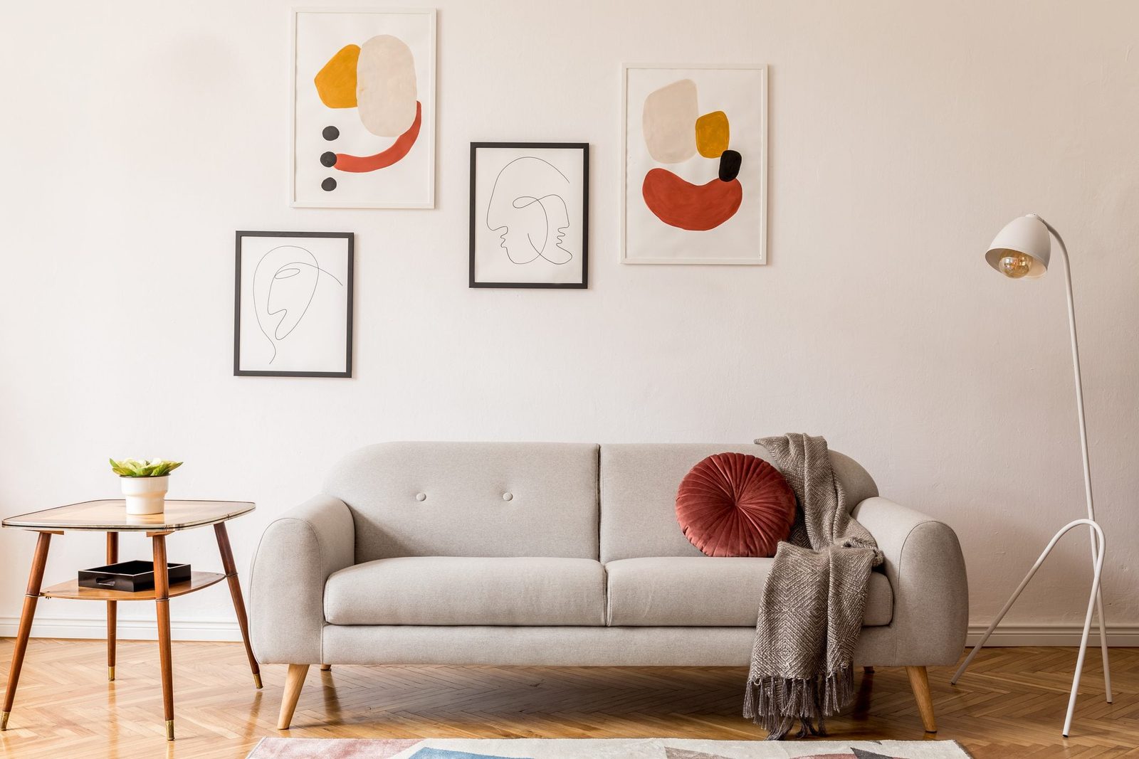 A light-colored sofa with a round pillow and a throw blanket sits beside a wooden table with a plant, under framed abstract art on a beige w