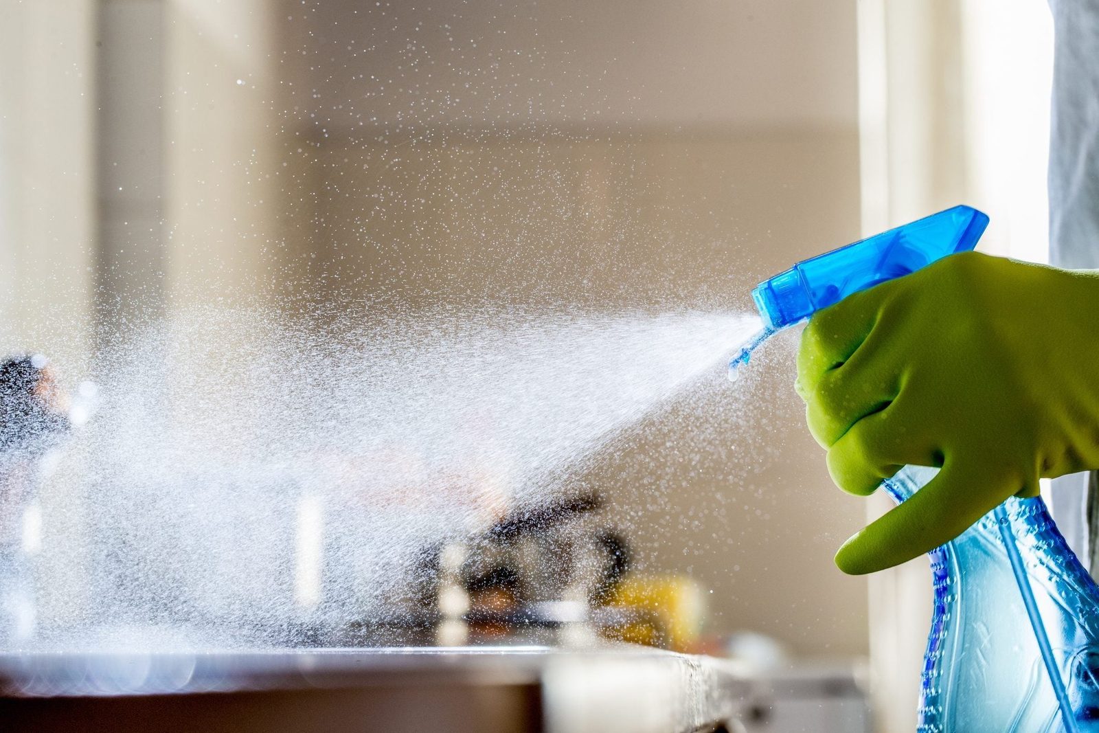 A gloved hand uses a spray bottle, releasing a fine mist of cleaning solution, while a blurred kitchen setting serves as the backdrop.