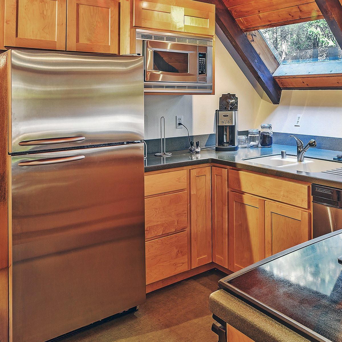 A stainless steel refrigerator stands in a modern kitchen, alongside a microwave and sink, with wooden cabinets and natural light from a sky