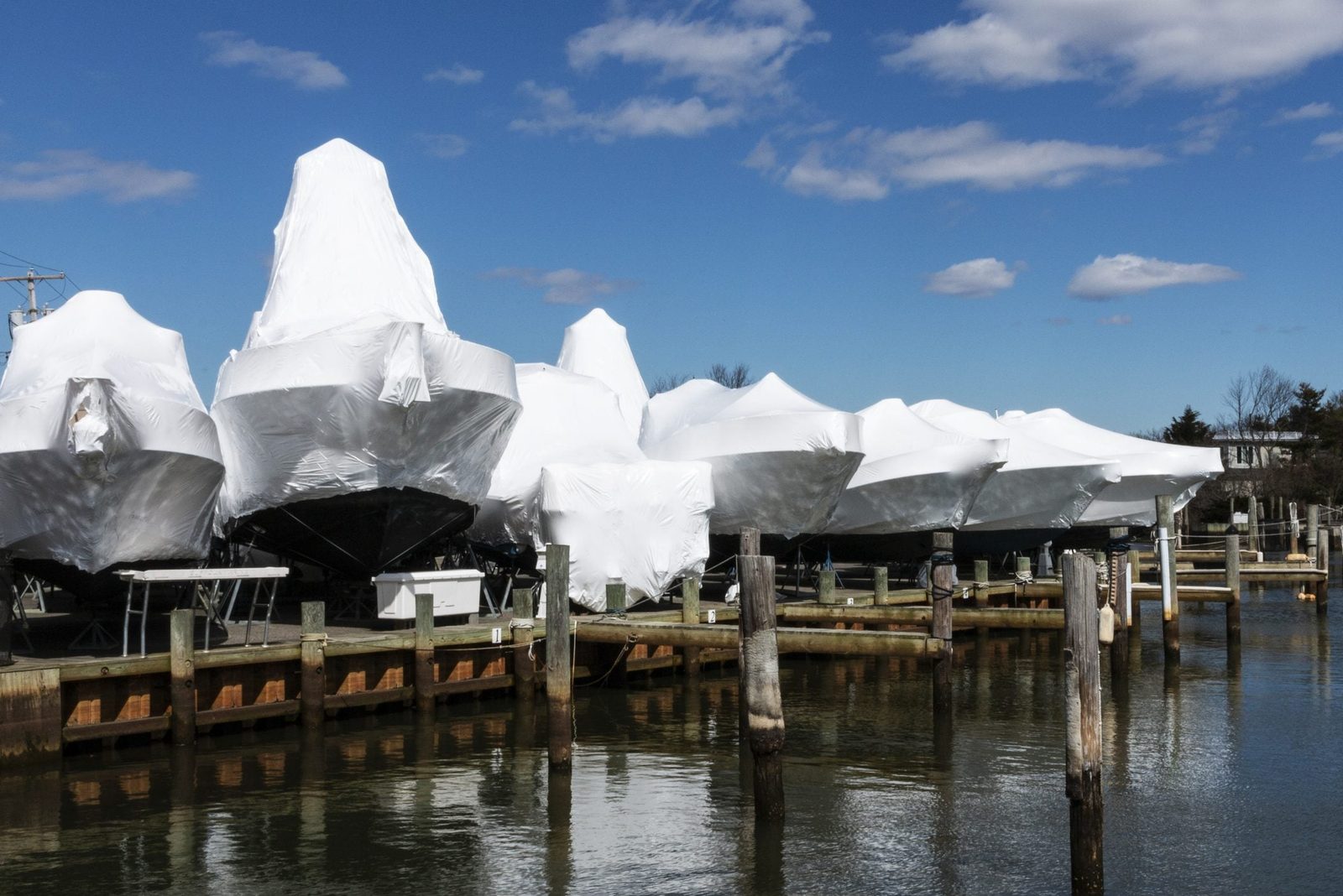 Boats wrapped in white tarps sit on a dock. They are stationary, surrounded by calm water under a blue sky with scattered clouds.
