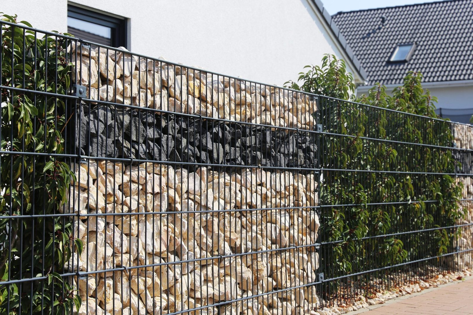 A wire fence supports stacked stones in distinct colors while green plants grow alongside. The setting features a modern building with a slo