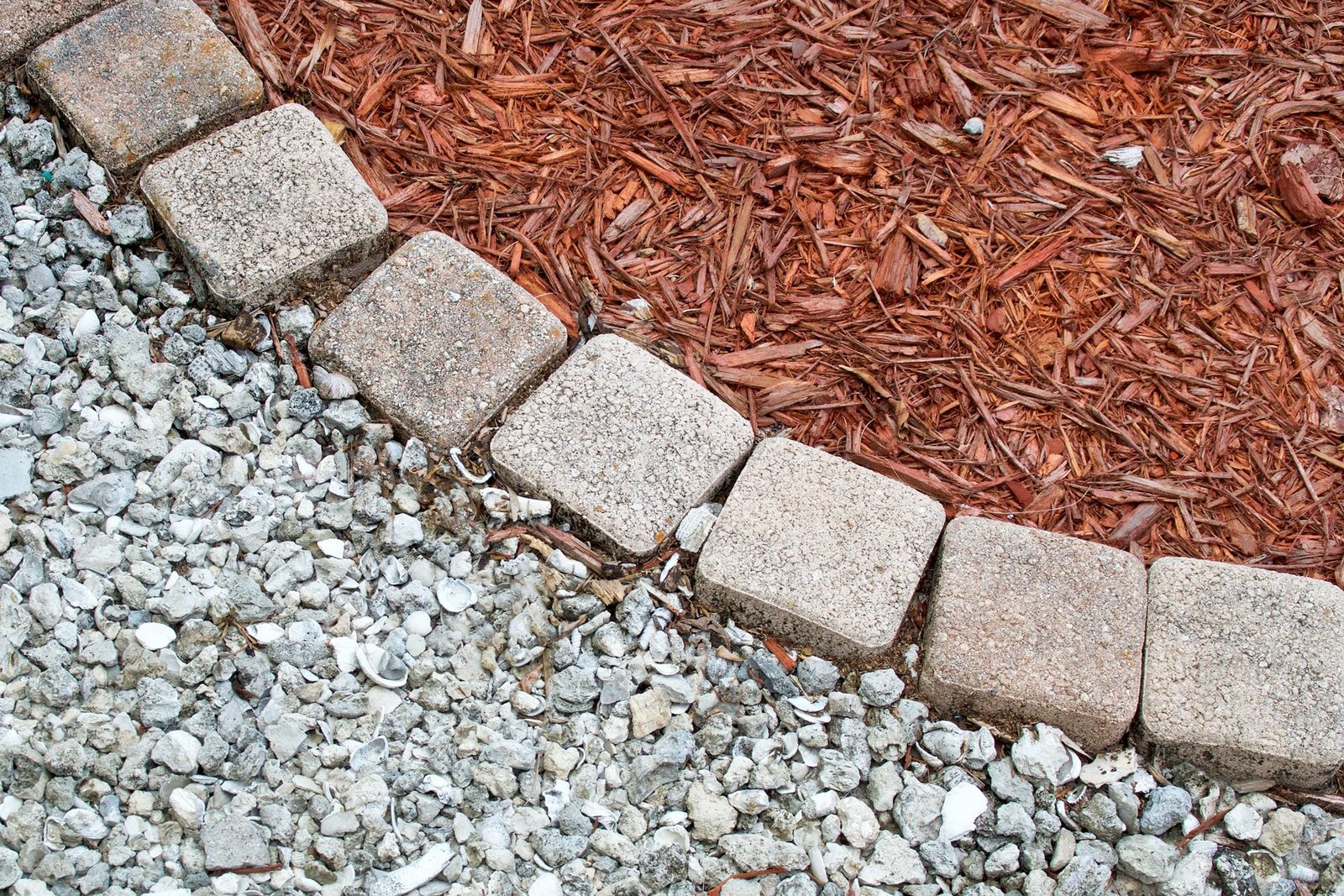 Concrete stones line the edge, separating a bed of reddish mulch from a coarse, pebble-strewn surface in an outdoor garden setting.