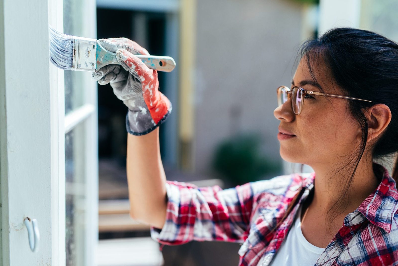 A woman paints a window frame with a brush while wearing gloves, surrounded by an outdoor area with soft lighting and blurred background ele