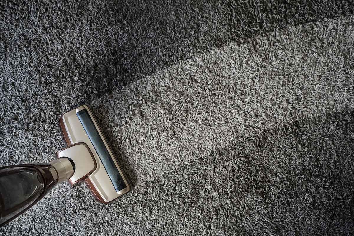 A vacuum cleaner glides across a textured carpet, creating a path of cleared fibers in a dimly lit room.