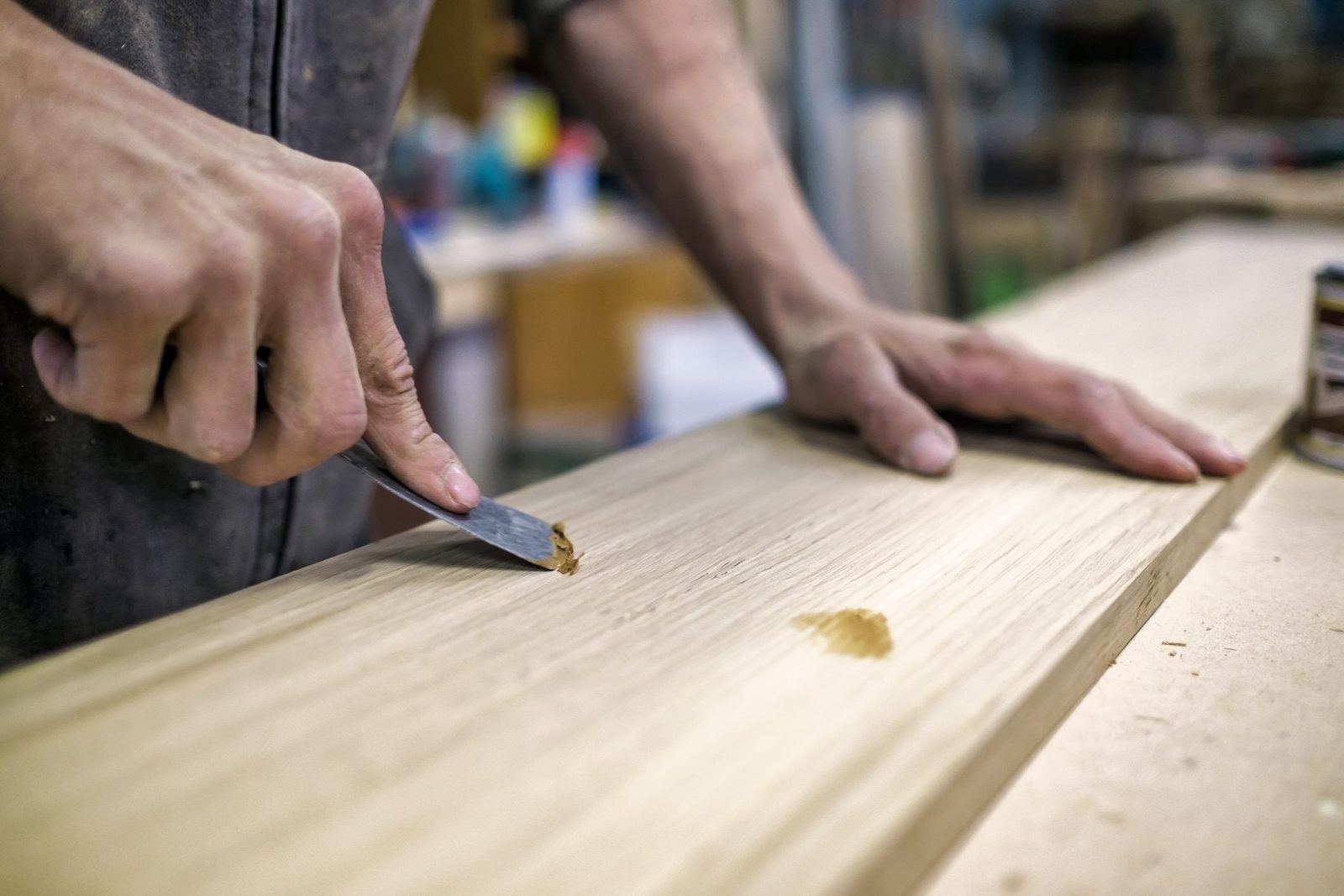 A hand uses a chisel to smooth a wooden surface in a workshop, with a can of finish nearby and other supplies blurred in the background.